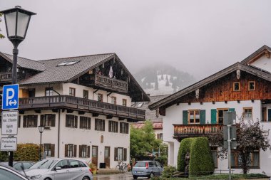 Oberammergau, Almanya - 14 Eylül 2024 - Geleneksel ahşap evleri, park edilmiş arabaları ve sislerle kaplı dağları olan sisli bir dağ köyü.