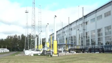 Sigulda, Letonya - 10 Ağustos 2024 - Metal yapıları, sarı tankları ve yüksek beyaz bacaları olan sanayi tesisleri, bulutlu bir gökyüzünün altında yeşil çimenler ve ağaçlarla çevrili.