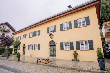 Bavyera, Almanya - 14 Eylül 2024 - Yeşil panjurlu sarı bir ev, kemerli kapı aralığı ve tuhaf bir Avrupa köyünde kaldırım taşı caddesi, bitkiler ve ön planda bir bank.