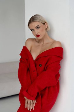 Kalın kırmızı rujlu ve sarı saçlı bir kadın, minimalist beyaz bir duvara yaslanmış omuz üstü kırmızı bir ceket giyiyor..