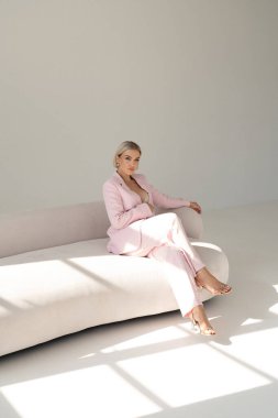 Pembe takım elbiseli kendine güvenen bir kadın minimalist bir ortamda, yumuşak doğal ışıkla yıkanmış modern beyaz bir kanepede oturuyor..