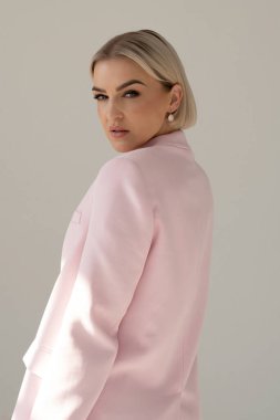 Pembe ceketli, kendine güvenen, sarı saçlı bir kadın omzunun üstünden bakar, zarafet ve tarz saçar yumuşak doğal ışıkla..