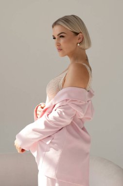 Pembe takım elbiseli kendine güvenen bir kadın ceketini örtüyor, dantelli bluzunu gösteriyor, yumuşak doğal ışıkta duruyor, zarif ve dengeli bir ifadeyle..