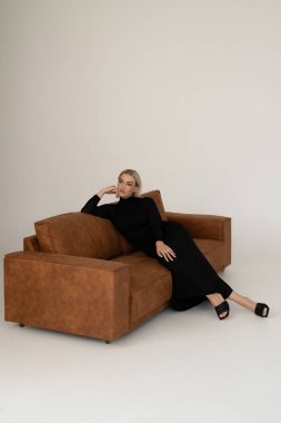 Pürüzsüz, siyah bir elbise giymiş bir kadın kahverengi bir kanepede uzanır. Minimalist, modern bir ortamda, tarafsız tonlarda, zarafet ve tarz yayar..