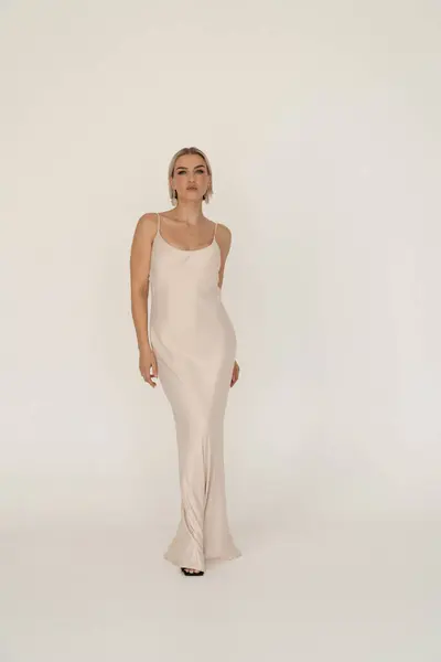 stock image A blonde woman stands confidently in a long beige dress, posing against a plain white background. She wears statement earrings, exuding elegance and style.