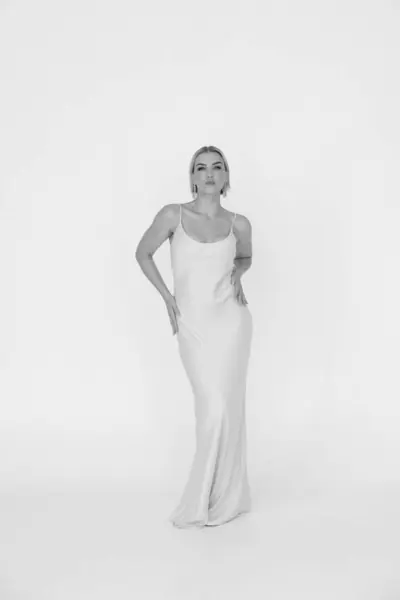 stock image A blonde woman stands confidently in a long beige dress, posing against a plain white background. She wears statement earrings, exuding elegance and style.