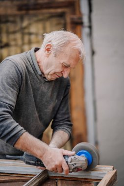 Yaşlı bir adam zımpara makinesiyle ahşap bir pencere çerçevesini zımparalamaya odaklanır. Açık hava atölyesinde marangozluk işine girişir..