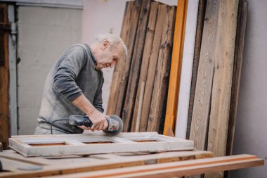 Yaşlı bir adam, çeşitli ahşap kalaslarla çevrili bir atölyedeki elektrikli aletle ahşap pencere çerçevesini zımparalıyor..