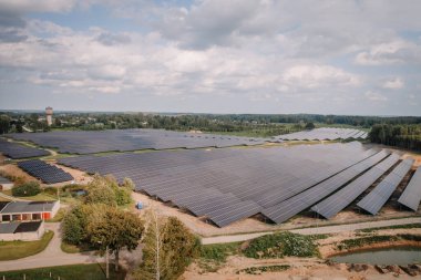 Kırsal bir bölgede parçalı bulutlu bir gökyüzünün altında sıra sıra güneş panelleri olan uçsuz bucaksız bir güneş çiftliğinin havadan görünüşü..