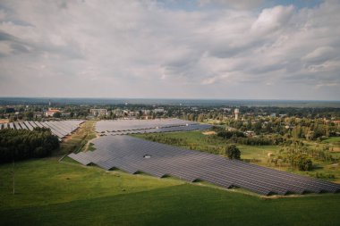 Yeşil alanlar ve uzak binalar arasında yenilenebilir enerji sağlayan, kırsal bir kasabanın yakınındaki sıra sıra güneş panelleri olan bir güneş çiftliğinin havadan görünüşü.