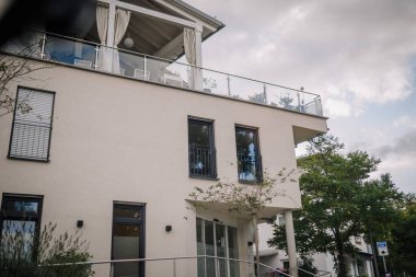 Bavaria, Germany - September 15, 2024 - Modern building with a large balcony and glass railing, surrounded by greenery under a cloudy sky. clipart