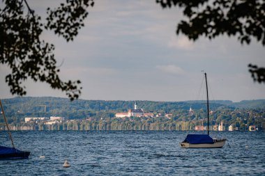 Bavyera, Almanya - 15 Eylül 2024 - Bir yelkenli sakin bir göl üzerinde yüzüyor, ağaç dalları tarafından çerçevelenmiş, uzak bir köy manzaralı ve tepe üzerinde büyük tarihi bir bina.