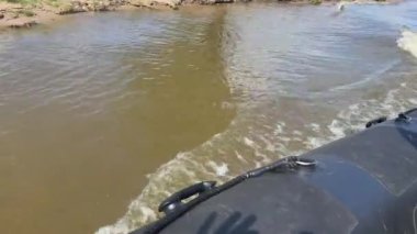 Kumlu bir nehir kıyısında seyreden lastik bir tekne güneşli bir günde ilerlerken suda küçük dalgalar yaratır..