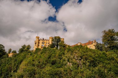 Bavyera, Almanya - 15 Eylül 2024 - Almanya 'nın Bavyera kentindeki bulutlu bir gökyüzünün altında yemyeşil bir tepenin üzerindeki Hohenschwangau Kalesi' nin çarpıcı manzarası.