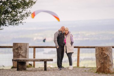 Bavyera, Almanya - 15 Eylül 2024 - Bir çift, arka planda uçan bir paraglider ile Bavyera 'da manzaralı bir bakış açısında poz veriyor, açık hava deneyiminin tadını çıkarıyor.