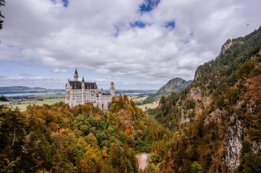 Bavyera, Almanya - 15 Eylül 2024 - Neuschwanstein Şatosu sonbahar yapraklarıyla çevrili, arka planda dağlar ve vadiler bulutlu bir gökyüzünün altında.