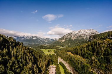 Tirol, Avusturya - 15 Eylül 2024 - Parlak mavi bir gökyüzü altında karla kaplı dağların, sık ormanların ve dolambaçlı yolların çarpıcı panoramik manzarası.