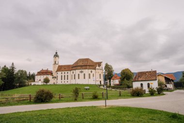 Bavyera, Almanya - 15 Eylül 2024 - Bulutlu bir günde çevresi yeşilliklerle çevrili büyük tarihi bir kilise binası, kırsal bir alanda küçük bir şapel ve evler.
