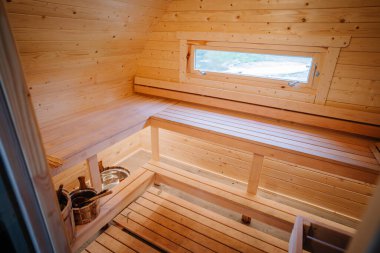 Sıcak, ahşap bir saunanın içinde banklar, küçük bir pencere ve geleneksel sauna aksesuarları var. Sıcak ve sakin bir atmosfer yaratıyorlar..