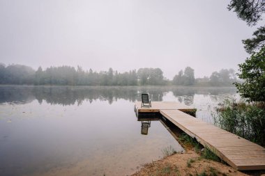 Sade bir göl manzarası, ahşap bir iskele, tek bir sandalye, ve sudaki sisli yansımalar, sakin bir doğal ortamda ağaçlarla çevrili..