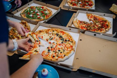 Riga, Letonya - 19 Eylül 2024 - Deniz ürünleri pizzasından bir dilim alan bir kişiyle birlikte kutularda birkaç pizza. Roka ve zeytin gibi farklı malzemeler görülebilir..