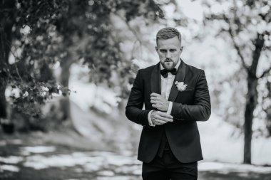 Valmiera, Letonya - 15 Ağustos 2024 - Kol düğmelerini ayarlayan, koyu renk takım elbiseli ve yaka çiçekli bir damat, açık hava düğününe hazırlanıyor