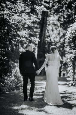 Valmiera, Letonya - 15 Ağustos 2024 - Bir gelin ve damat bir orman yolunda el ele yürür, düğün günlerinde samimi bir anı paylaşırlar.