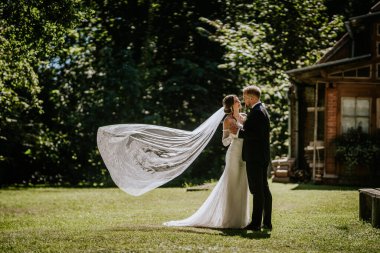 Valmiera, Letonya - 15 Ağustos 2024 - Gelin ve damat açık havada romantik bir anı paylaşıyor, örtüsü rüzgarda zarifçe süzülürken, yemyeşil ve arka planda kırsal bir ev.