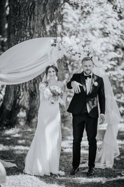 Valmiera, Letonya - 15 Ağustos 2024 - Neşeli bir gelin ve damat seremoniden sonra yüzüklerini gösterir, bir çiçek kemerinin altında durur, açık havada siyah ve beyaz bir sette yakalanırlar..