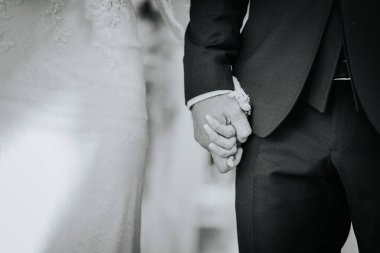 Valmiera, Letonya - 15 Ağustos 2024 - Düğün törenlerinde resmi kıyafetlerini ve yüzüklerini sergileyen gelin ve damadın el ele tutuşurken çekilmiş siyah beyaz fotoğrafı.