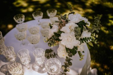 Kristal gözlüklü bir masa seti ve beyaz bir çiçek aranjmanı, açık hava kutlaması için hazır, güneş ışığı ve gölgeler zarafeti artırıyor..