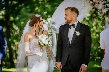 Valmiera, Letonya - 15 Ağustos 2024 - Neşeli bir gelin ve damat arka planda çiçek kemeri ve yeşillik olan açık hava düğün töreninde el ele tutuşup gülümsüyorlar..