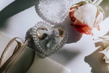 Valmiera, Letonya - 15 Ağustos 2024 - Kalp şeklinde bir kristal tutacağa bağlanmış iki altın alyansın yakın çekimi, masada bir gülün yanında, aşkı simgeliyor..