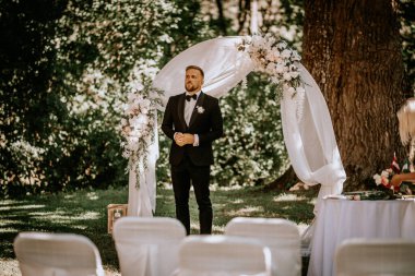 Valmiera, Letonya - 15 Ağustos 2024 - Siyah smokinli bir damat, arka planda yeşillik olan açık hava seremonisinde çiçekli bir düğün kemerinin altında duruyor.