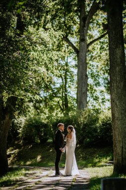 Valmiera, Letonya - 15 Ağustos 2024 - Bir gelin ve damat, düğün günlerinde yemyeşil yemyeşil ile çevrili, gün ışığında, ağaçlarla kaplı bir yolda el ele dururlar..