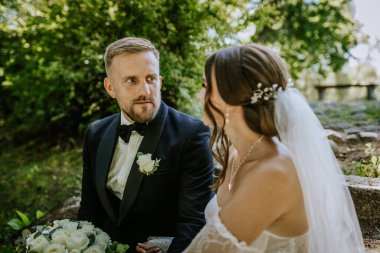 Valmiera, Letonya - 15 Ağustos 2024 - Bir gelin ve damat açık havada oturur, birbirine sevgiyle bakar, arka planda yeşillik, hassas bir düğün anını yakalar.