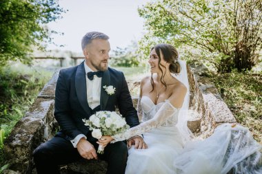 Valmiera, Letonya - 15 Ağustos 2024 - Gülümseyen bir gelin ve damat açık havada el ele tutuşarak ve bir buket beyaz gülle düğün günlerinde neşeli bir anı paylaşıyorlar..