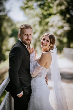 Valmiera, Letonya - 15 Ağustos 2024 - Güneşli bir günde gelin ve damat birlikte poz verir, gelin damadın elini nazikçe tutar, kameraya bakar, zarafet saçar