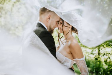 Valmiera, Letonya - 15 Ağustos 2024 - Bir gelin ve damat akıcı bir peçenin altında, alınları birbirine değmiş, yumuşak doğal bir ışıkla çevrili, açık hava ortamında.