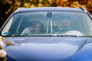 Valmiera, Letonya - 15 Ağustos 2024 - Gelin, damat ile bir arabada otururken, ön camdan gülümseyerek, düğünlerinden sonra uzaklaşmaya hazır bir barış işareti gösterir..