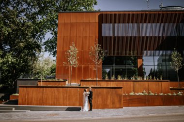 Valmiera, Latvia - August 15, 2024 - A bride and groom stand in front of a large modern rust-colored building, sharing a romantic moment on their wedding day. clipart