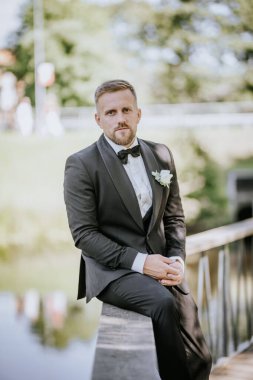 Valmiera, Letonya - 15 Ağustos 2024 - Bir damat ahşap bir korkulukta düşünceli bir şekilde oturur, siyah bir smokin giyer, arka planda sakin bir açık hava ortamı vardır..