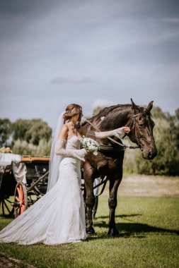 Valmiera, Letonya - 15 Ağustos 2024 - Beyaz gelinlikli bir gelin bir buket tutuyor ve güneşli bir günde açık havada at arabası ile geziyor..