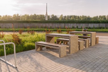 Valmiera, Latvia - August 8, 2024 -  Outdoor stone picnic table and benches set on a paved area with green grass and bushes in the background under soft sunlight. clipart