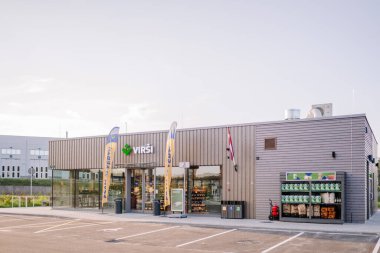 Valmiera, Latvia - August 8, 2024 - A VIRSI convenience store with large windows, flags, and an outdoor product display featuring firewood, charcoal, and windshield washer fluid. clipart