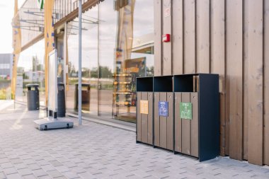 Valmiera, Letonya - 8 Ağustos 2024 - Temiz bir çevrede geniş pencereli ve ahşap dış görünüşlü modern bir mağazanın önünde plastik, kağıt ve cam için geri dönüşüm kutuları.