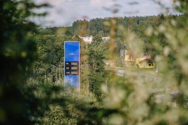 Valmiera, Letonya - 8 Ağustos 2024 - Çevresi kırsal yol ve binalarla çevrili bir benzin istasyonu fiyat tabelası.