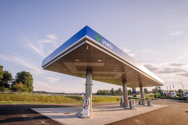 Valmiera, Letonya - 8 Ağustos 2024 - Temiz bir gökyüzü altında yakıt pompaları olan modern bir benzin istasyonu, çevresindeki yeşillikler ve uzak mesafede görülebilen bir kamyon durağı.