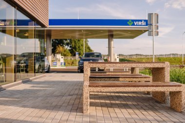 Valmiera, Letonya - 8 Ağustos 2024 - Güneşli bir günde park halindeki bir benzin istasyonunun yanındaki taştan bankta ve camlı bir binada.