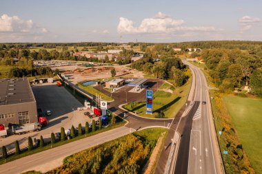 Valmiera, Letonya - 8 Ağustos 2024 - Parçalı bulutlu bir gökyüzü altında kamyonları, binaları ve manzaralı kırsal çevresi olan bir otoyol boyunca uzanan bir benzin istasyonunun havadan görünüşü.